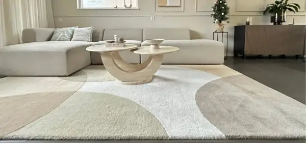 Ein Wohnzimmer mit einem beigen Ecksofa, einem runden Couchtisch und einem modernen Teppich im geometrischen Design. Dieser Teppich-Trend bringt Ruhe und Stil in jeden Raum.