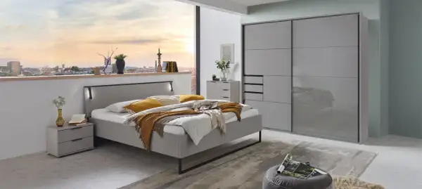 Modernes Schlafzimmer mit grauem Bett, Nachttischen und einem großen Kleiderschrank. Ein Fenster gibt den Blick auf eine Stadt bei Sonnenuntergang frei.