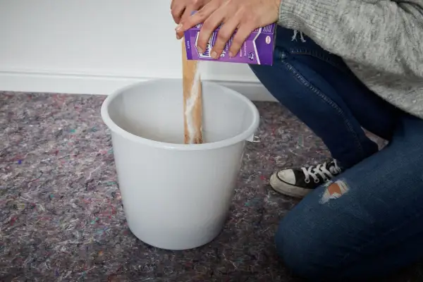Hände schütten Pulver aus einer Packung in einen Eimer, um Tapetenkleister aufzutragen.