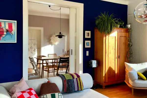 Ein Wohnzimmer mit einer tiefblauen Wand, einem Holzkasten, Pflanzen und einem gemütlichen Sofa. Durch eine Tür ist ein Esszimmer mit Holztisch und Stühlen zu sehen.